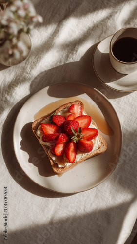 Aesthetic breakfast scene with white plate featuring toast topped with cream and fresh strawberries, accompanied by cup of coffee on sunlit table