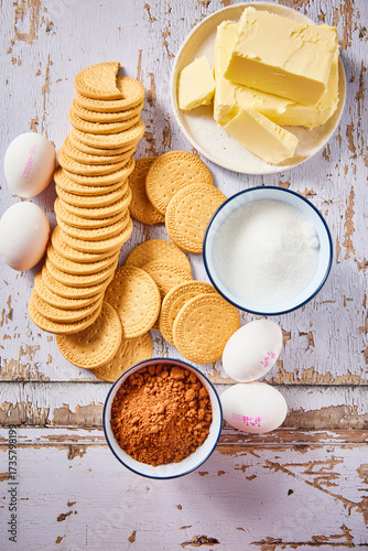 Flat lay of ingredients for traditional Dutch arretjescake, also known as refrigerator cake. Butter, cocoa powder, sugar, eggs, and biscuits arranged on a rustic wooden background.