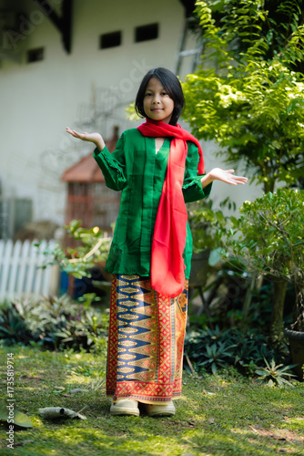 Young girl wearing Indonesian traditional dress with green kebaya and red scarf, smiling outdoors in cultural fashion portrait.