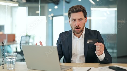 Worried handsome businessman in a professional suit experiences a shocking online payment issue with a credit card on a laptop at an office desk, showing frustration.