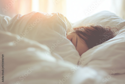 Young person sleeping peacefully under soft white blankets in a sunlit bedroom, cozy morning scene with warm natural light creating a calm and comfortable atmosphere.