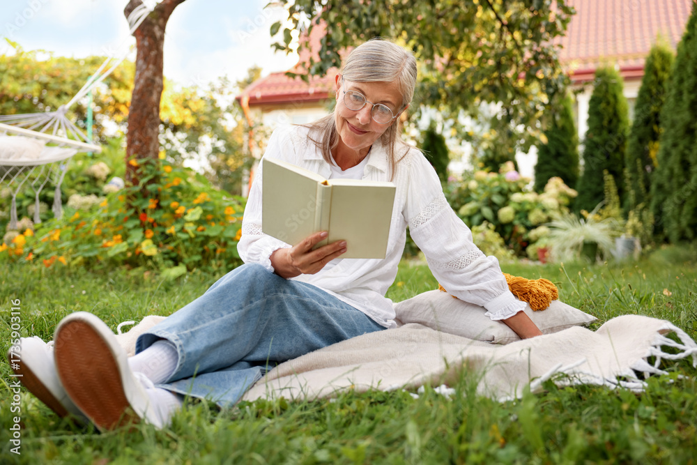 Fototapeta premium Senior woman reading book on blanket in garden