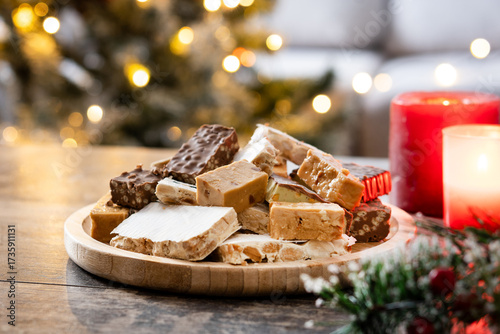 Assorted traditional Christmas nougat on wooden plate and christmas tree background