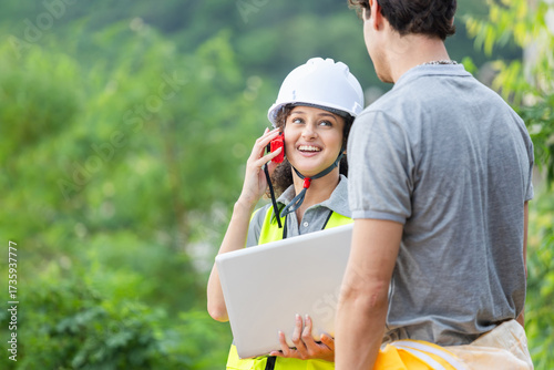 Smiling Female Engineer Communicating with Walkie Talkie on Site, Construction Manager Using Laptop and Radio for Team Coordination, Happy Project Supervisor Discussing Work Outdoors with Colleague