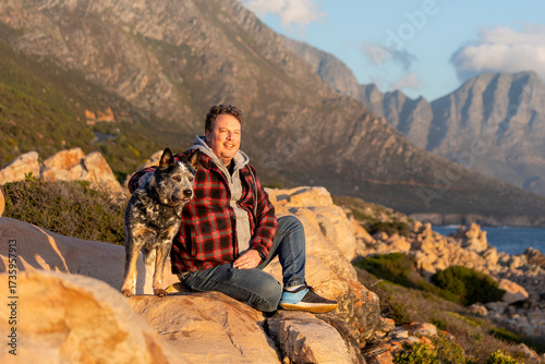 A man sitting outdoors with his Australian Cattle dog (blue heeler), looking at the view, hiking and exploring together, the dog is alert