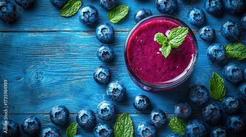 Blueberry smoothie in glass surrounded by fresh blueberries and mint on blue wooden background.