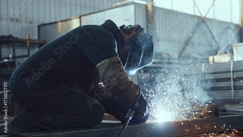 Skilled worker in protective gear actively performing welding work, generating bright sparks and smoke while fabricating metal components in a manufacturing environment