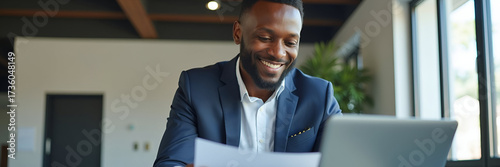 Happy busy young African business man checks documents while working on laptop in office smiles as businessman accountant company employee reads law paper corporate finances invoice report concept as 