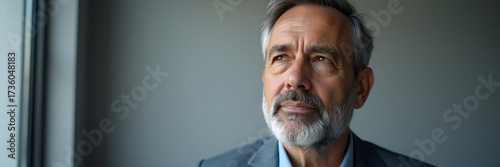 A confident middle-aged man gazes thoughtfully away at a gray office wall, embodying inspiration and ambition; accompanied by generous copy space to insert text or logos.