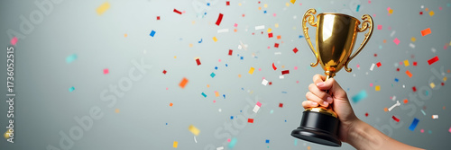 A hand holding a large gold trophy adorned with intricate details, surrounded by colorful confetti on a soft grey background, emphasizing success and victory. Ample copy space available.