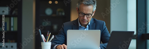 A serious, middle-aged businessman focuses intently on a laptop, seated at an office desk immersed in investment project details, suggesting authority and professionalism with ample copy space.