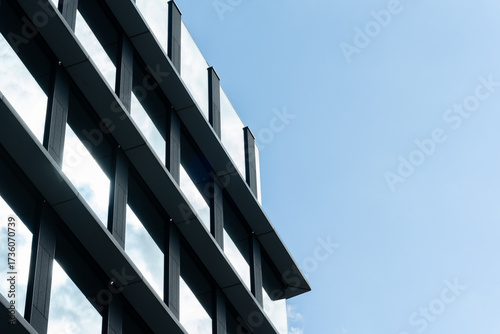 Contemporary glass and steel office building facade with geometric lines and clear blue sky background. For illustrating architecture, business, or urban development concepts.