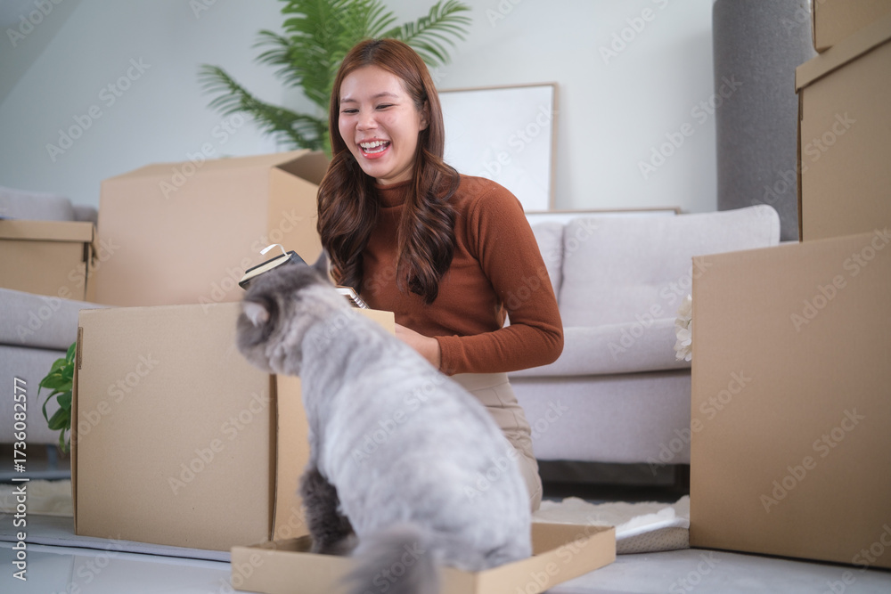 Fototapeta premium Happy woman laughs as her cat plays in a box during unpacking.