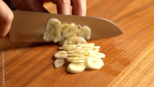A close-up of a hand slicing garlic into thin rounds on a wooden cutting board.