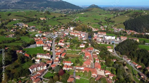 Aerial drone view of Lierganes, a historic village in Cantabria, Spain. The scene shows traditional stone houses, red-tiled rooftops, the medieval bridge over the Miera River, and lush green hills