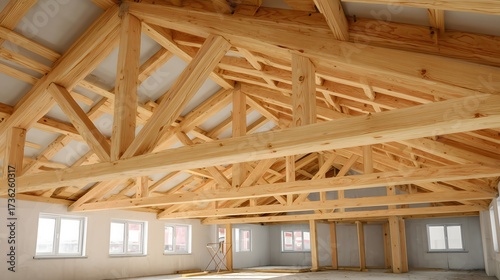 Interior view of a spacious room with wooden beams and trusses. Large windows allow natural light to fill the space. The walls are unfinished, showcasing raw materials.