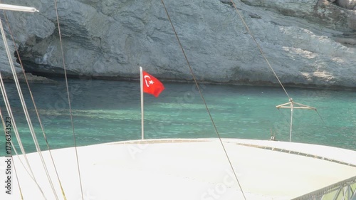 A tranquil scene showing the bow of a boat anchored near massive, sunlit rocky cliffs. The Turkish flag is visible in the frame, adding a sense of location and pride. 