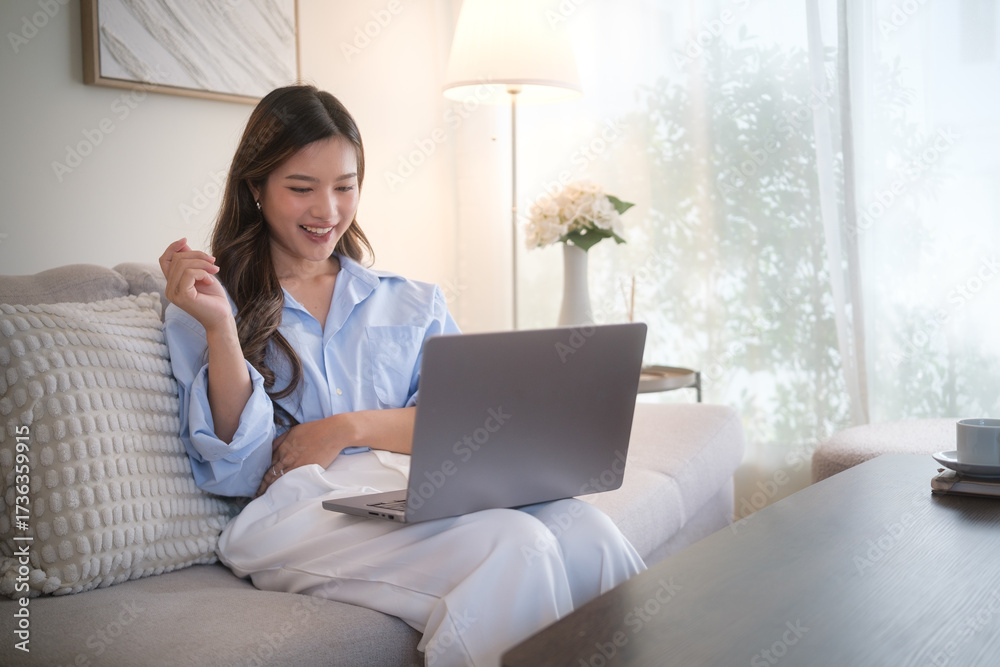 Fototapeta premium Young woman working from home on her laptop with a joyful smile in a cozy, modern living room.