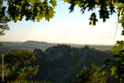 Photography Scenic view of massive rock formations surrounded by dense forest, framed by green leaves in warm sunlight, symbolizing wilderness, timeless nature, and tranquility