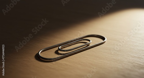 of a metallic paperclip on a light brown surface, illuminated by directional lighting which creates a stark contrast of light and shadow, presenting a minimalist and simple composition.