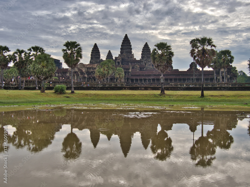 Obraz premium Angkor Wat Temple Reflection - The iconic spires of Angkor Wat reflected in a tranquil pool, surrounded by palm trees under a dramatic, cloudy sky.