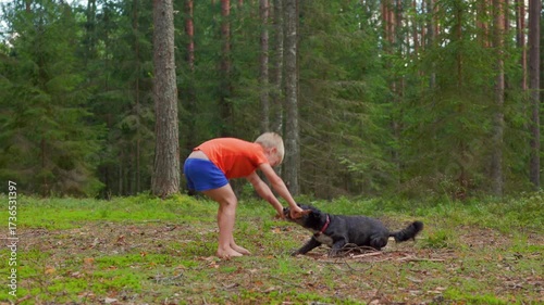 In slow motion, a barefoot boy in orange shirt plays with a black dog in a sunny forest clearing, showing joy, energy, and childhood fun