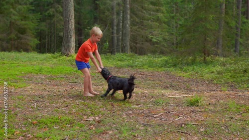 In slow motion, a barefoot boy in orange shirt plays with a black dog in a sunny forest clearing, showing joy, energy, and childhood fun