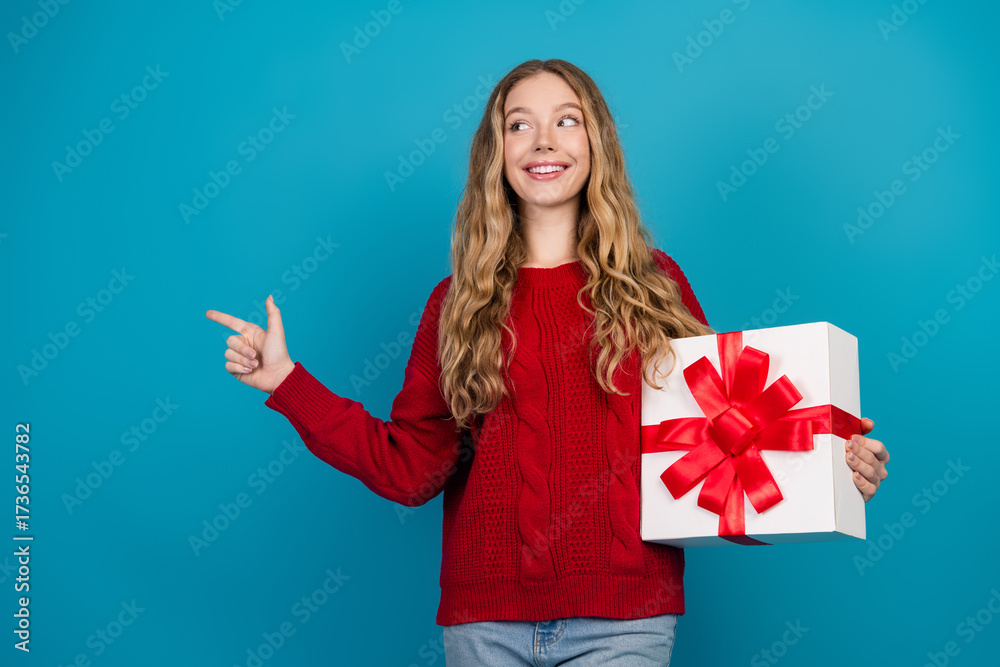 Fototapeta premium Adorable young woman in a red sweater holding a gift box wrapped with a red bow against a vibrant blue background