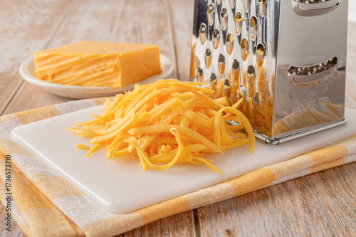 Grated cheddar cheese close-up. Shredded orange cheese and grater on a white cutting board over rustic wooden tabletop. Dairy product, calcium concept.