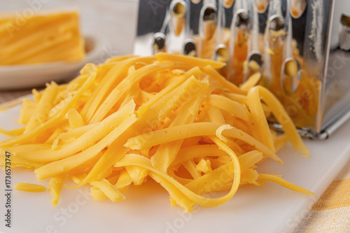 Shredded cheddar cheese close-up. Heap of grated orange cheese and metal grater on a white cutting board. Dairy product, milk protein, calcium concept.