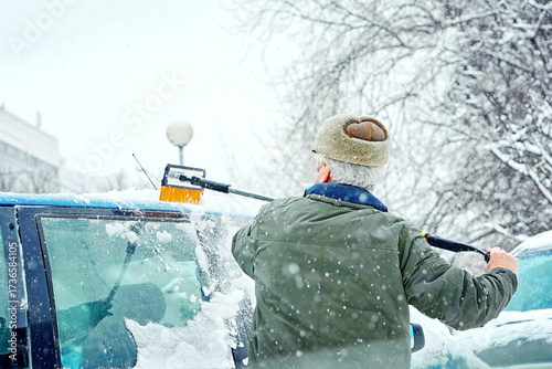 Senior man uses brush to clean snow off car during heavy snow storm, ensuring visibility and safety in harsh winter weather conditions