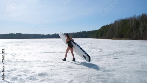 Woman in black swimsuit holding sup board walking on frozen ice snow on river lake. Happy model in warm boots. Brunette female enjoy sunny day on beautiful nature with forest, outdoor recreation.