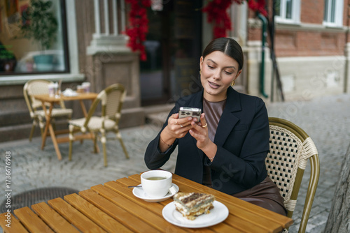 Stylish woman using her smartphone at an outdoor cafe with tea and cake. Dressed in a black blazer and chic outfit, she captures moments in a charming urban setting.
