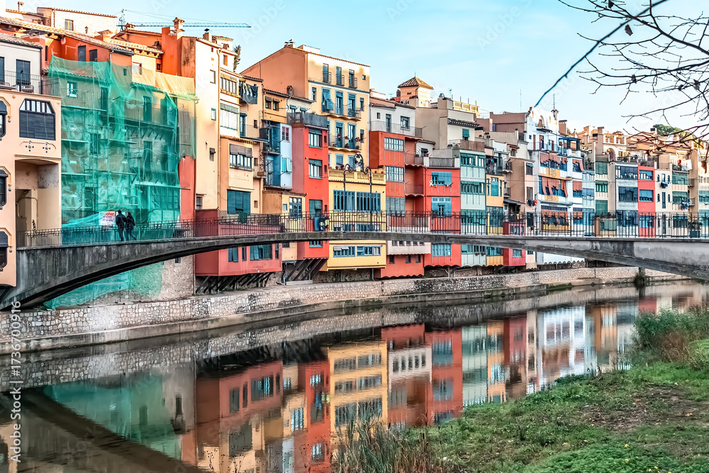 Fototapeta premium Colorful riverside houses with reflections in the Onyar River and Gomez Bridge, showcasing vibrant Spanish architecture. Girona, Spain - November 28, 2021