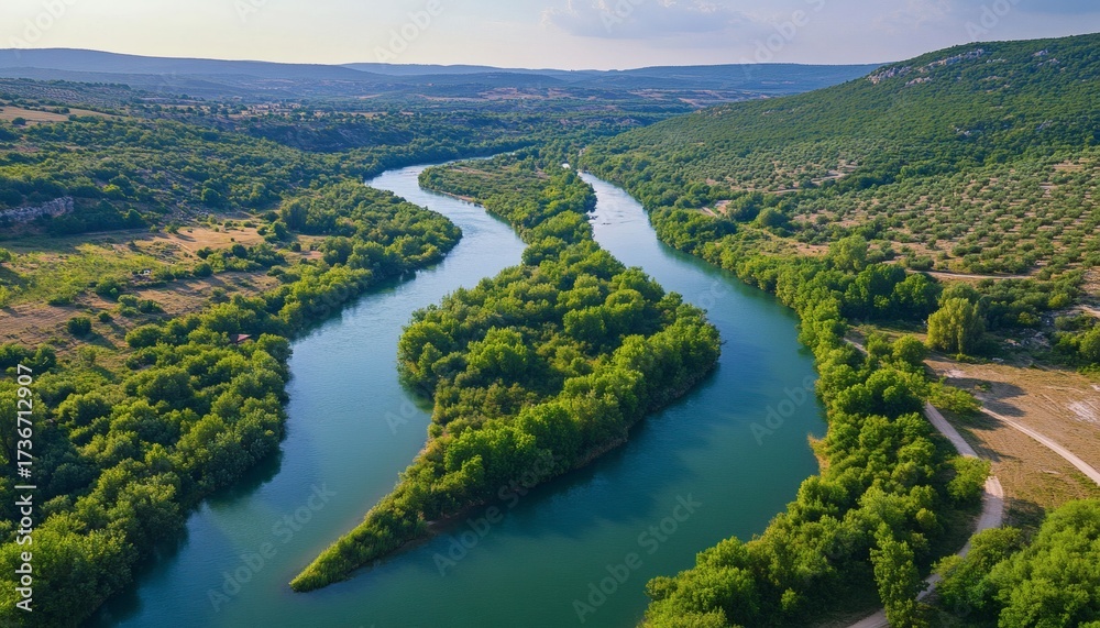 Obraz premium The image shows a river winding its way through a valley, surrounded by dense green vegetation and hills in the background.