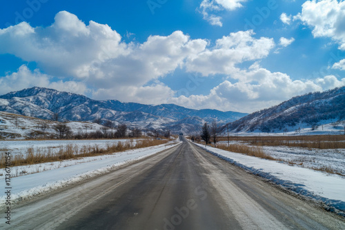 A long, clear road stretches through a snowy landscape, surrounded by majestic mountains and a bright blue sky filled with clouds