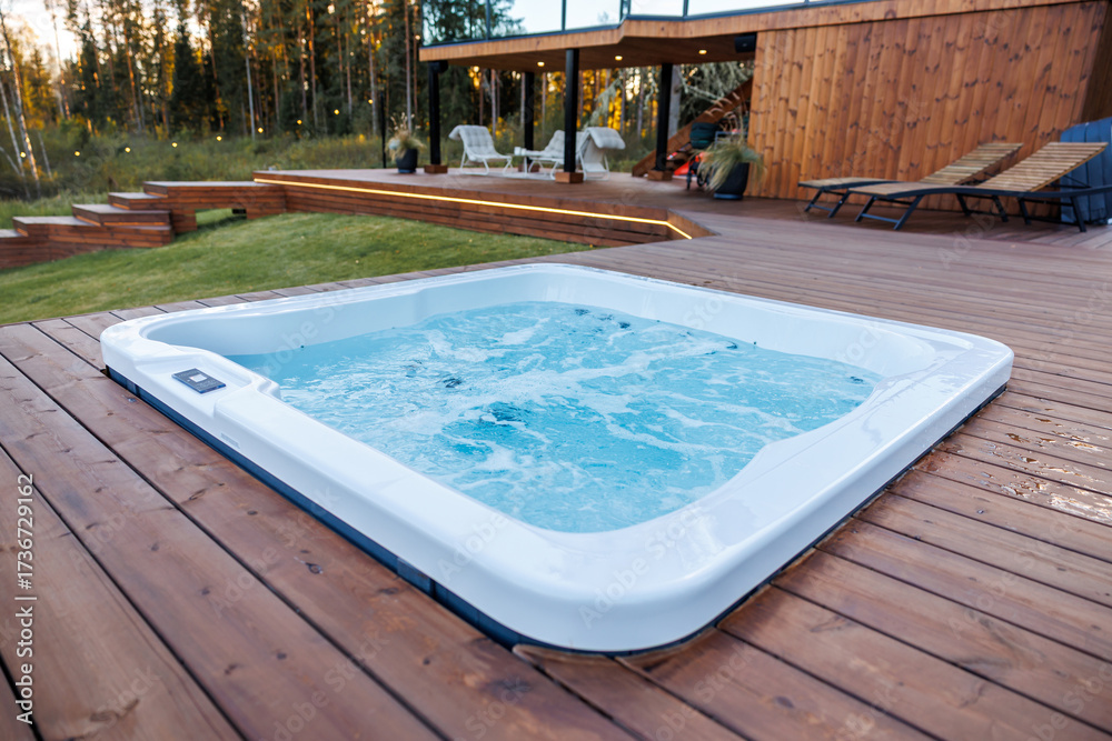 Obraz premium Modern hot tub set into a polished wooden deck with bubbling water, lounge chairs, string lights, and a wooden facade structure surrounded by tall trees.
