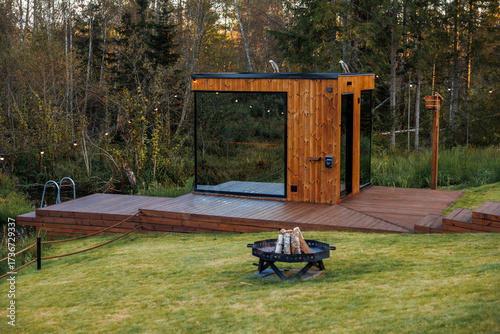 Cuadro en lienzo A modern wooden sauna with glass panels sits on a raised deck with string lights, a fire pit, a water feature, and surrounded by grassy areas and trees