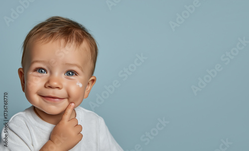 A cute smiling baby uses one finger to apply the cream himself on light blue background