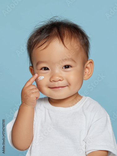 A cute japanese smiling baby uses one finger to apply the cream himself on light blue background