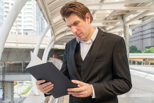 Businessman in a black suit reading documents outdoors in a modern city background. Concept of career, corporate work, planning, decision-making, and professional lifestyle.