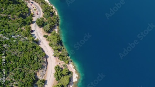 Drone footage captures a scenic aerial view of a winding coastal road nestled amidst lush green hills, bordering the vibrant turquoise waters of the Mediterranean Sea near Icmeler, Turkey.