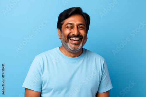 Happy Indian man smiling against blue background wearing light blue t-shirt. Portrait representing positivity, men's health awareness and support for Movember and November Blue.