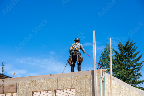 New housing development construction project, carpenter on wall in safety harness and hardhat, using pneumatic nail gun, work in progress
