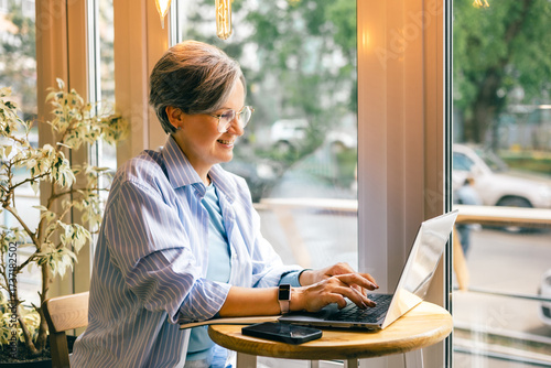 Senior woman smiling while working on laptop at cozy cafe table by the window with natural daylight