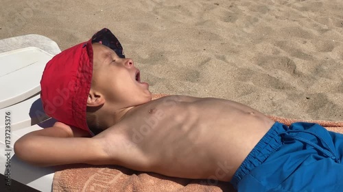 A young boy blissfully relaxes in the sun, eyes closed with a happy smile, traces of sand on his face, enjoying a peaceful moment outdoors.