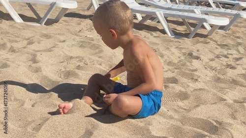 A young boy in blue swim trunks sits calmly on a sun-drenched beach, engaging with the sand as he gazes to the side, surrounded by lounging chairs.