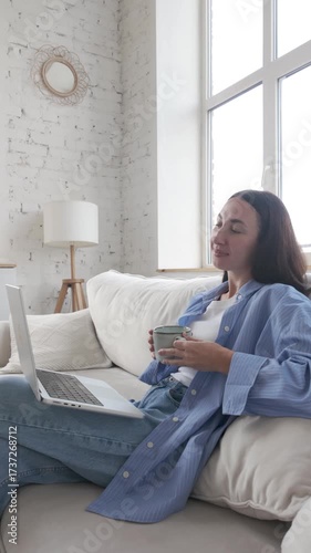 Brunette young woman in casual outfit relaxes on a sofa at home, holding a cup of coffee or tea while watching laptop. Cozy leisure time with calm mood and soft daylight