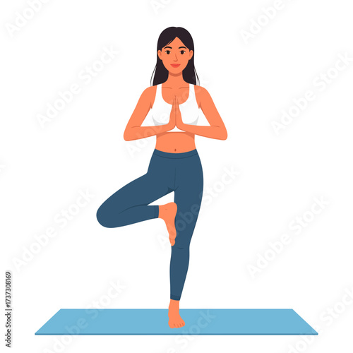 A young woman with dark hair performs a tree pose (Vrksasana) in yoga, balancing on one leg with hands in Anjali Mudra, wearing a white sports bra and blue leggings on a light blue mat.