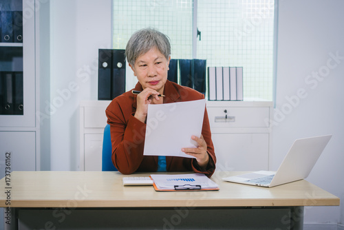 Senior Asian woman with professional office experience, focusing on reviewing financial reports and investment documents, reflecting her expertise in business, finance, and investment strategy